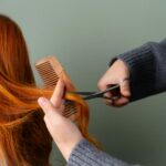 Close-up of red hair being trimmed with scissors and comb on green background.